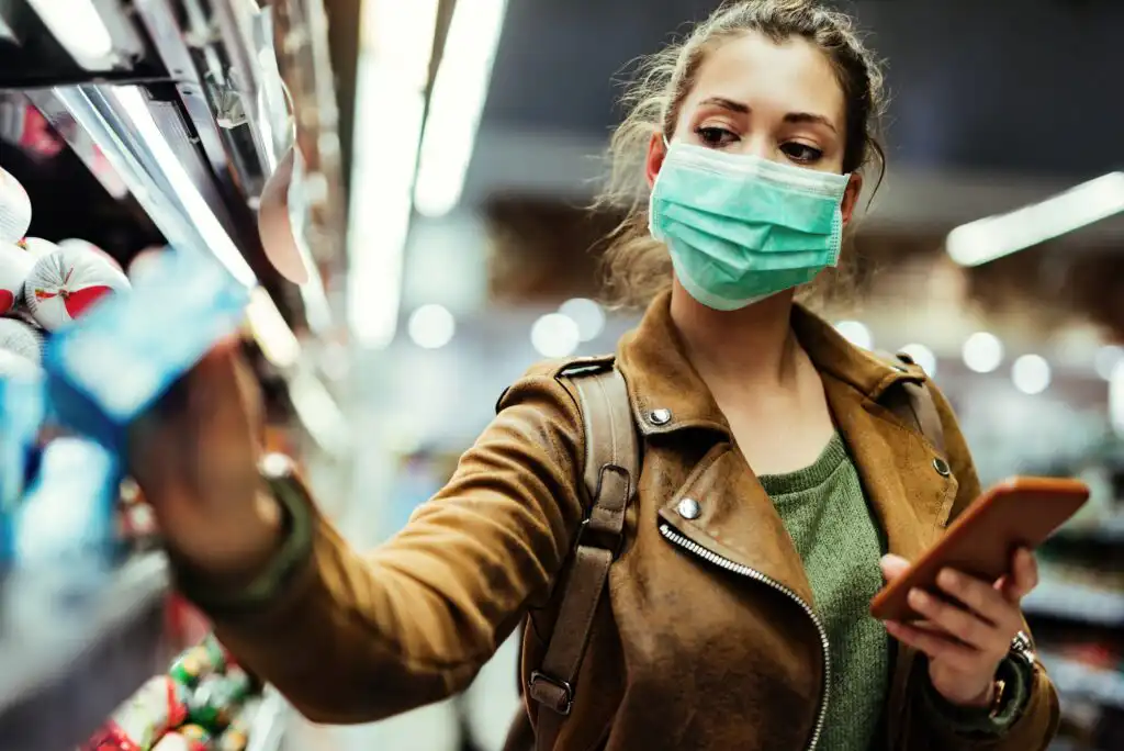 A woman wearing a face mask, brown jacket, and backpack holds a smartphone while reaching for an item on a grocery store shelf. She appears focused, shopping in a brightly lit aisle.