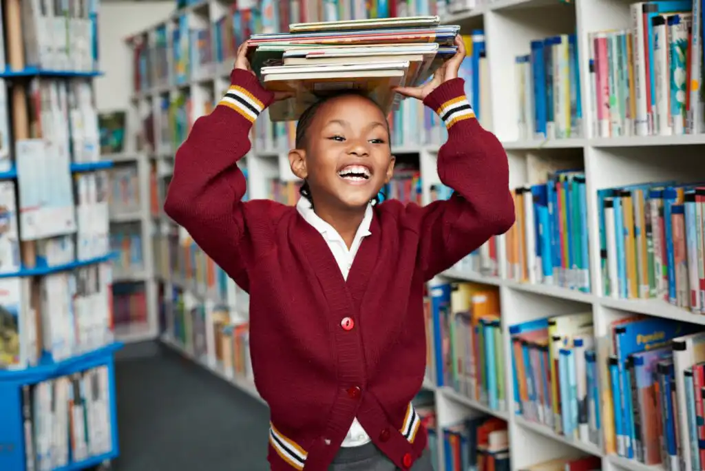 Smiling child in a red sweater balances a stack of books on their head while standing in a brightly lit library aisle filled with colorful bookshelves.
