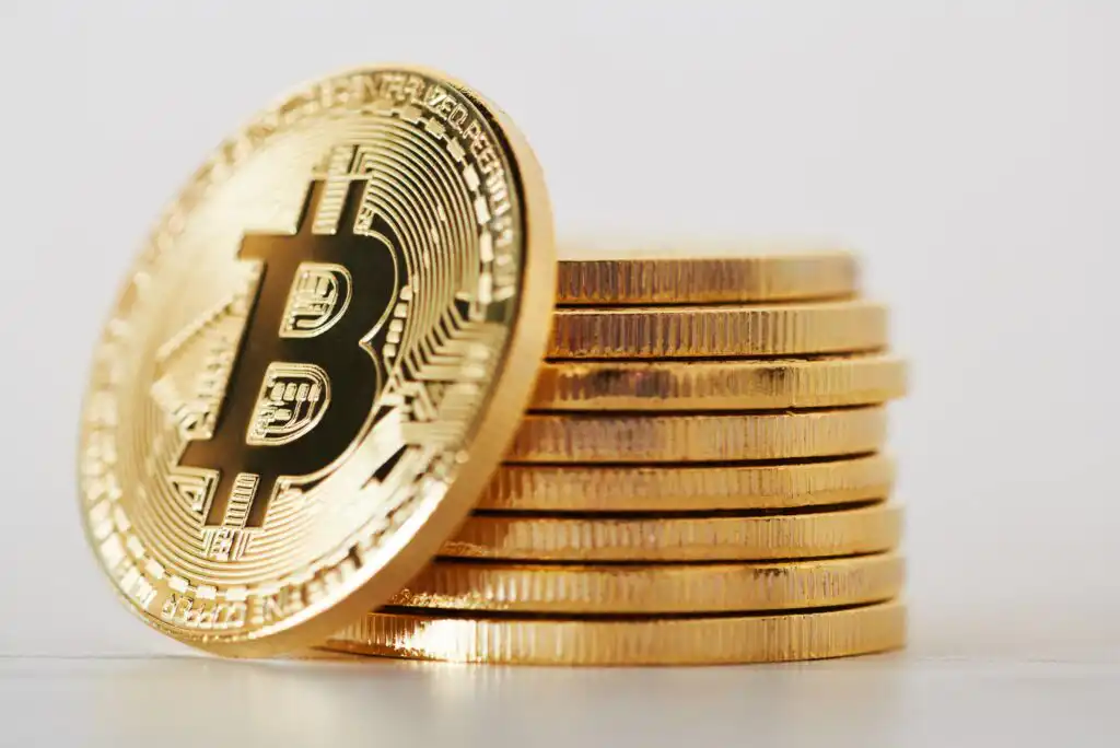 A stack of shiny gold coins with a prominent Bitcoin symbol on the front coin, set against a plain, light background.