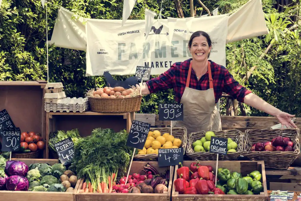A smiling vendor in an apron stands behind a farmers market stall filled with fresh produce like peppers, carrots, potatoes, beetroots, eggs, and cabbages. Signs with prices and a market banner are visible in the background.