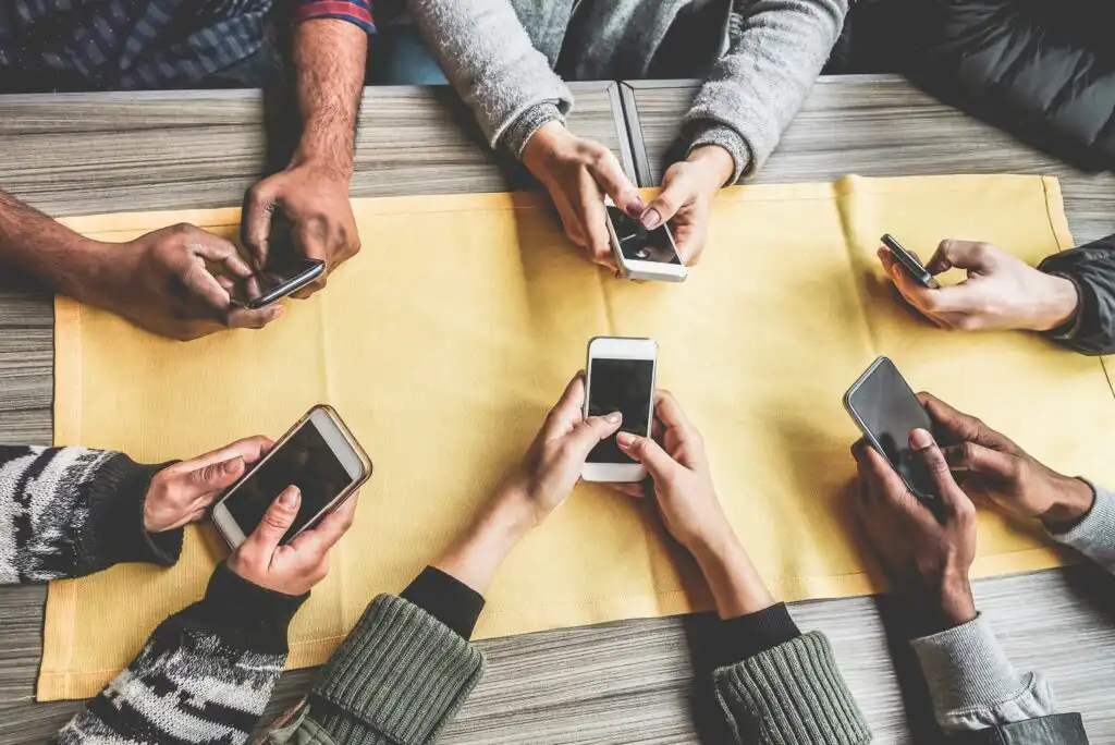 Six people sit around a table with a yellow runner, each holding and looking at their smartphones. Only their hands and phones are visible, indicating a group setting focused on mobile devices.