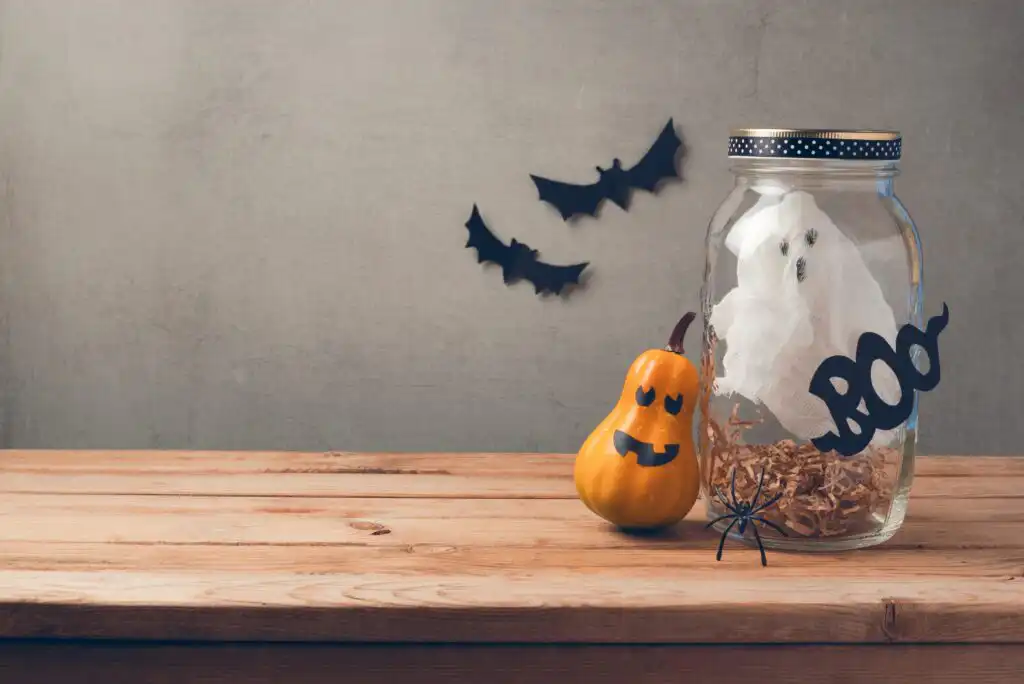 A Halloween scene with a jar holding a tissue ghost labeled “BOO,” a smiling pumpkin decoration, a plastic spider, and black paper bats on a plain wall, all arranged on a wooden surface.