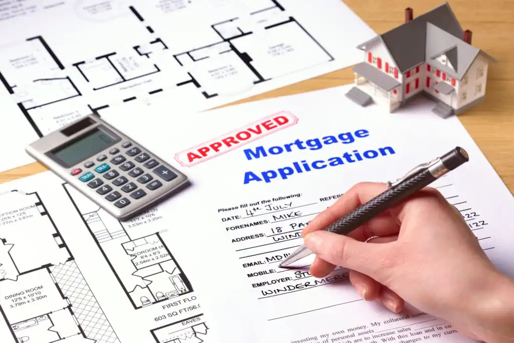 A hand fills out a mortgage application form stamped “APPROVED,” surrounded by architectural floor plans, a calculator, and a small model house.