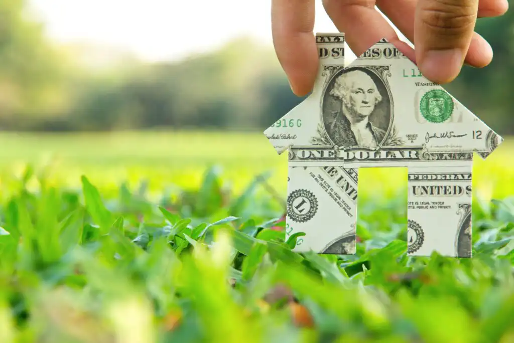 A hand holds a paper house made from a U.S. dollar bill, set against green grass and a blurred natural background.