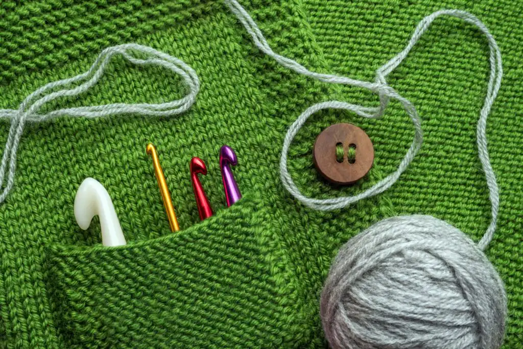 Green knitted fabric with a pocket holding four colorful crochet hooks, a large wooden button, a ball of gray yarn, and a gray strand of yarn arranged on top.