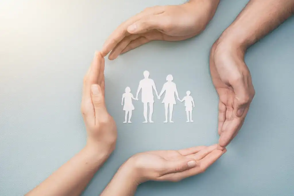 Four hands gently form a protective circle around a paper cutout of a family, consisting of two adults and two children, symbolizing care, safety, and protection.