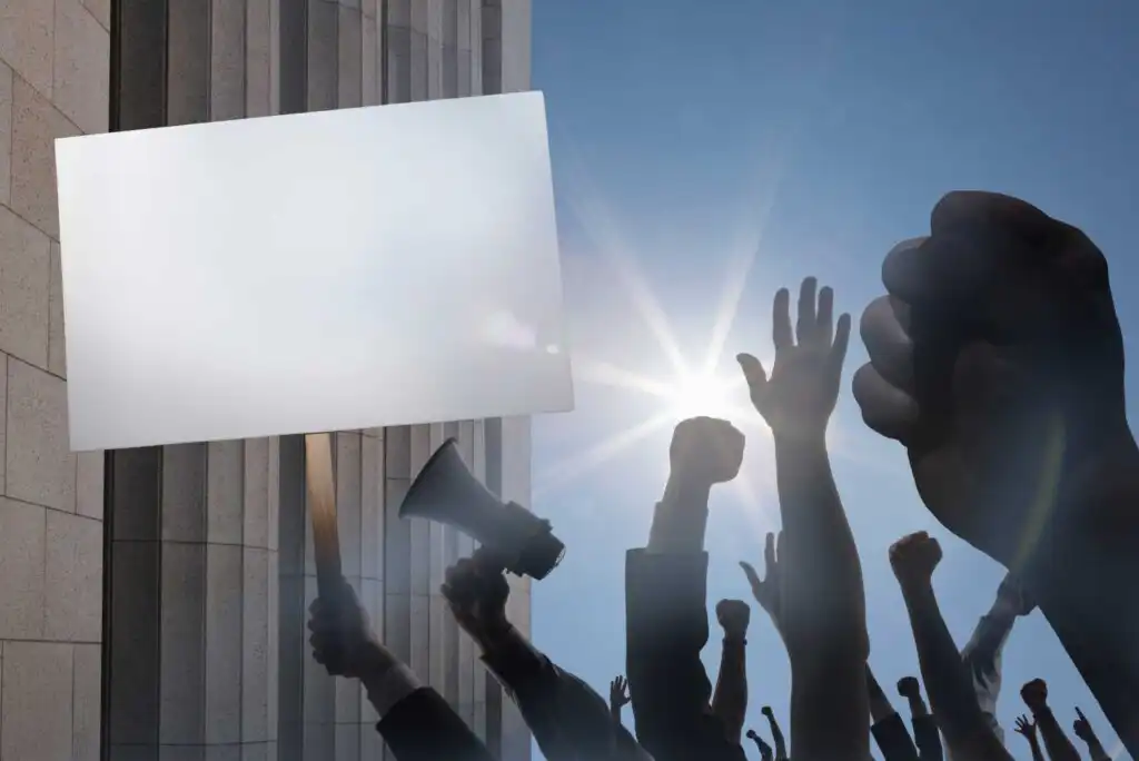A crowd of people protest outside a building, raising their fists and holding up a blank sign and a megaphone under a bright blue sky with the sun shining.