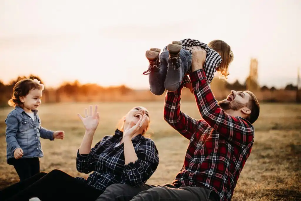 A cheerful family enjoys time outdoors at sunset; a man lifts a young child playfully, while a woman laughs and another child stands nearby, all smiling and dressed in casual clothes.