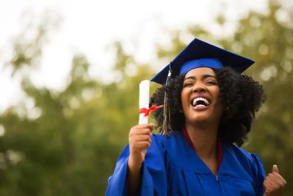 A smiling graduate in a blue cap and gown holds up a diploma tied with a red ribbon, celebrating outdoors with green trees in the background.