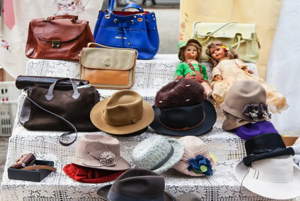 A display of vintage hats, handbags, and two dolls on lace-covered steps. The hats vary in color and style, while the bags are brown, blue, and cream. The dolls wear old-fashioned clothes and sit among the accessories.