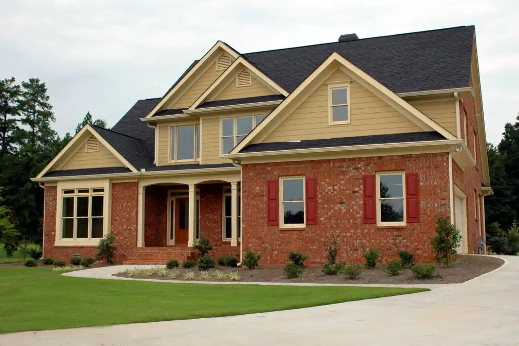 A two-story suburban house with tan siding, red brick exterior, dark shingles, and several large windows. There is a covered front porch, a curved driveway, and a well-kept lawn with small shrubs.