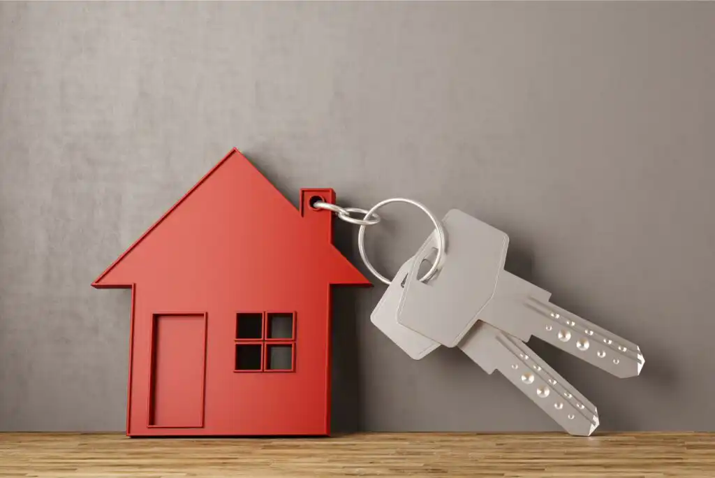 A red house-shaped keychain and two silver keys rest on a wooden surface against a plain gray background.