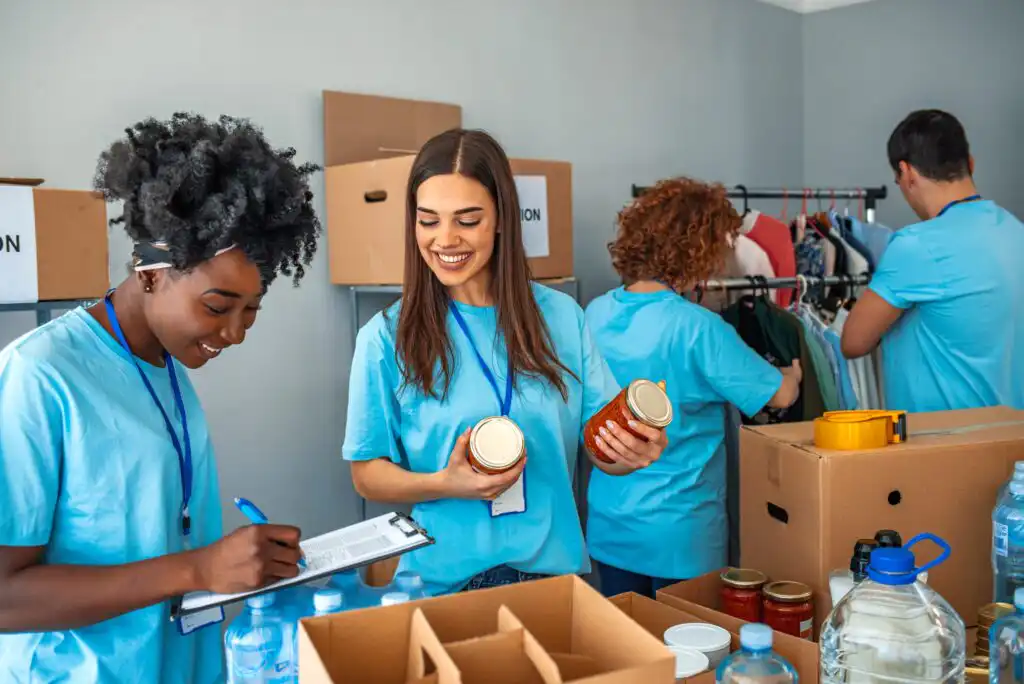 Four people wearing blue shirts volunteer at a donation center, sorting food items and clothing into boxes. Two women focus on canned goods, while another woman writes on a clipboard and a man hangs clothes.