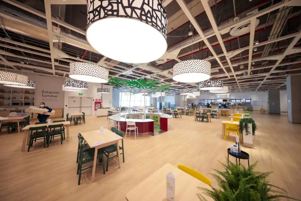 A spacious, modern cafeteria with wooden floors, colorful tables and chairs, large decorative ceiling lights, plants, and a person cleaning. The area is well-lit and mostly empty, creating an open and inviting atmosphere.