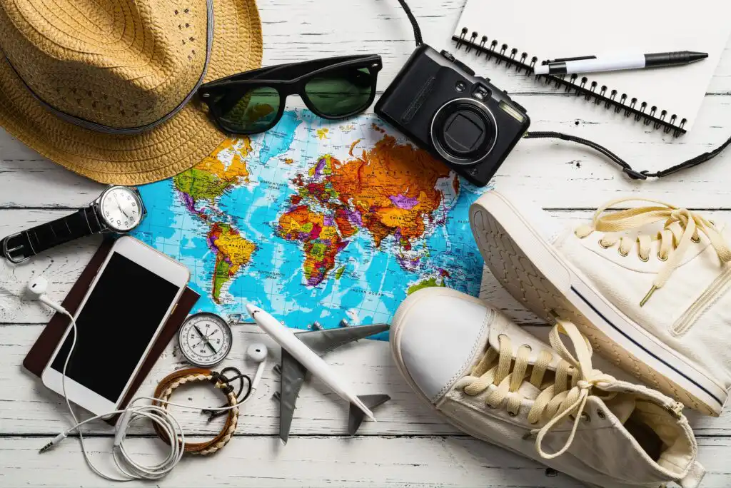 A travel-themed flat lay with a world map, straw hat, sunglasses, camera, notebook, pen, wristwatch, smartphone, earphones, compass, toy airplane, and white sneakers on a wooden surface.