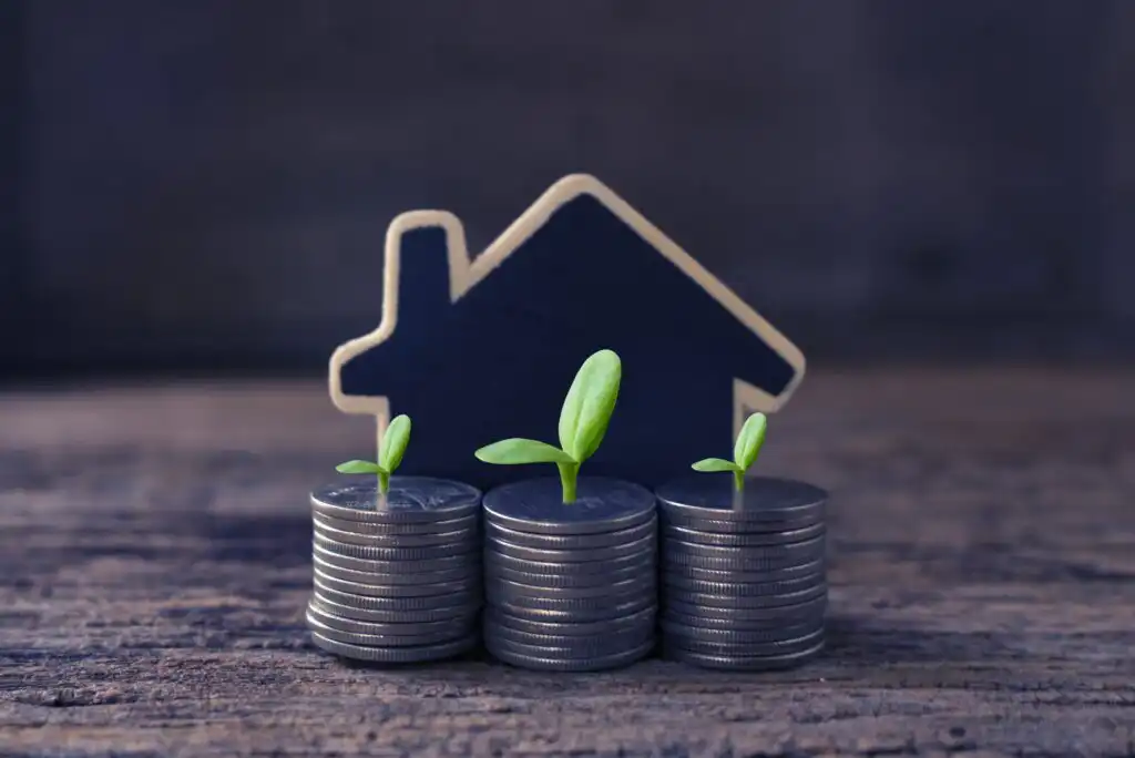 Three stacks of coins with small green plants growing from each stack, set in front of a house-shaped cutout, symbolizing investment growth and real estate savings.