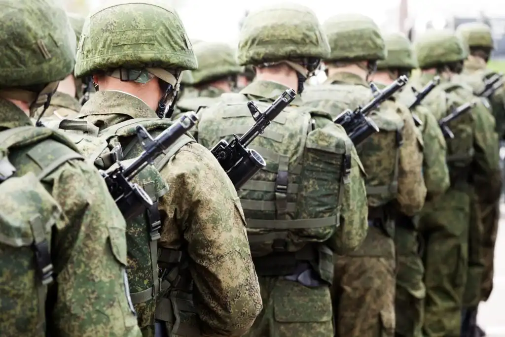 A group of soldiers in camouflage uniforms and helmets stand in formation with rifles slung across their backs, facing away from the camera.
