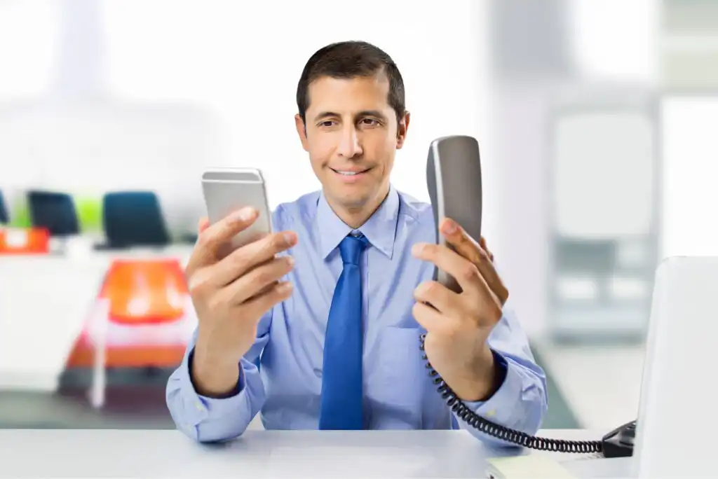 A man in a blue shirt and tie sits at a desk, smiling while holding a smartphone in one hand and a corded office phone in the other. An open laptop is on the desk. The background is an office setting.