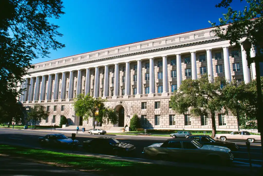 A large neoclassical government building with tall columns and multiple windows, facing a street with parked and moving cars, trees, and people walking on the sidewalk under a clear blue sky.