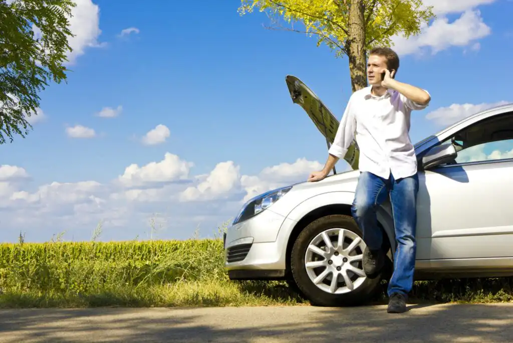 A man in a white shirt and jeans leans against a silver car with its hood open, talking on a cellphone by a rural road under a blue sky with clouds and trees in the background.
