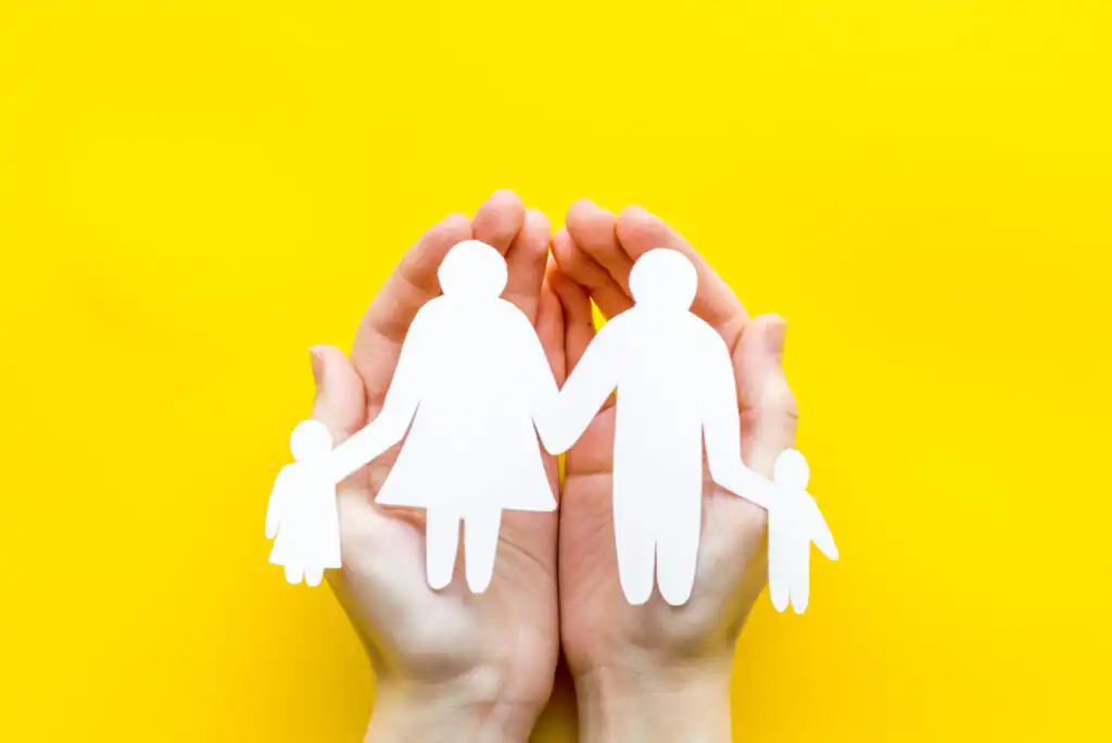 Two hands gently cup a paper cutout of a family with two adults and two children, all holding hands, against a bright yellow background.