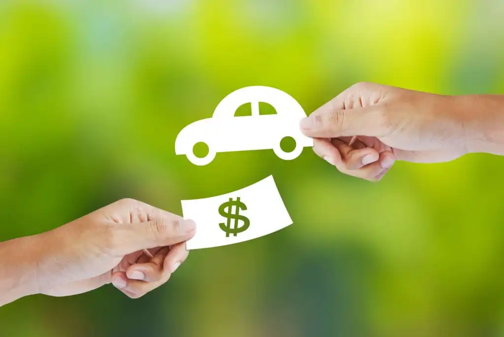 Two hands exchange paper cutouts, one of a car and the other of a dollar bill symbol, against a blurred green background, representing buying or selling a vehicle.