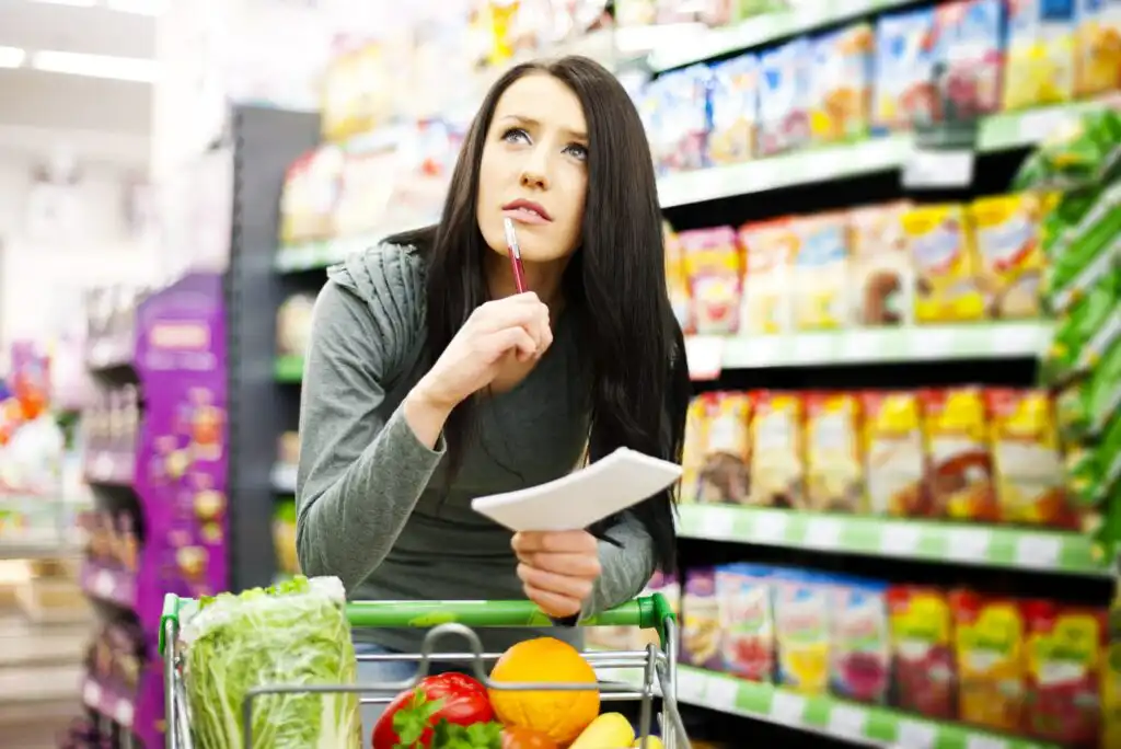 A woman in a grocery store leans on her shopping cart, holding a shopping list and pen, looking thoughtful. The cart contains fresh produce, and shelves filled with colorful packaged goods are visible in the background.