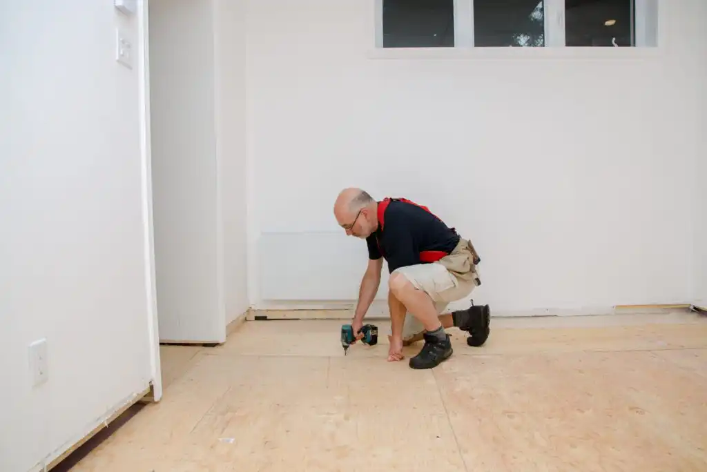 A person wearing glasses and work clothes kneels on a plywood floor, using a power drill to secure the floor in a bright, unfinished room with white walls and a small window.