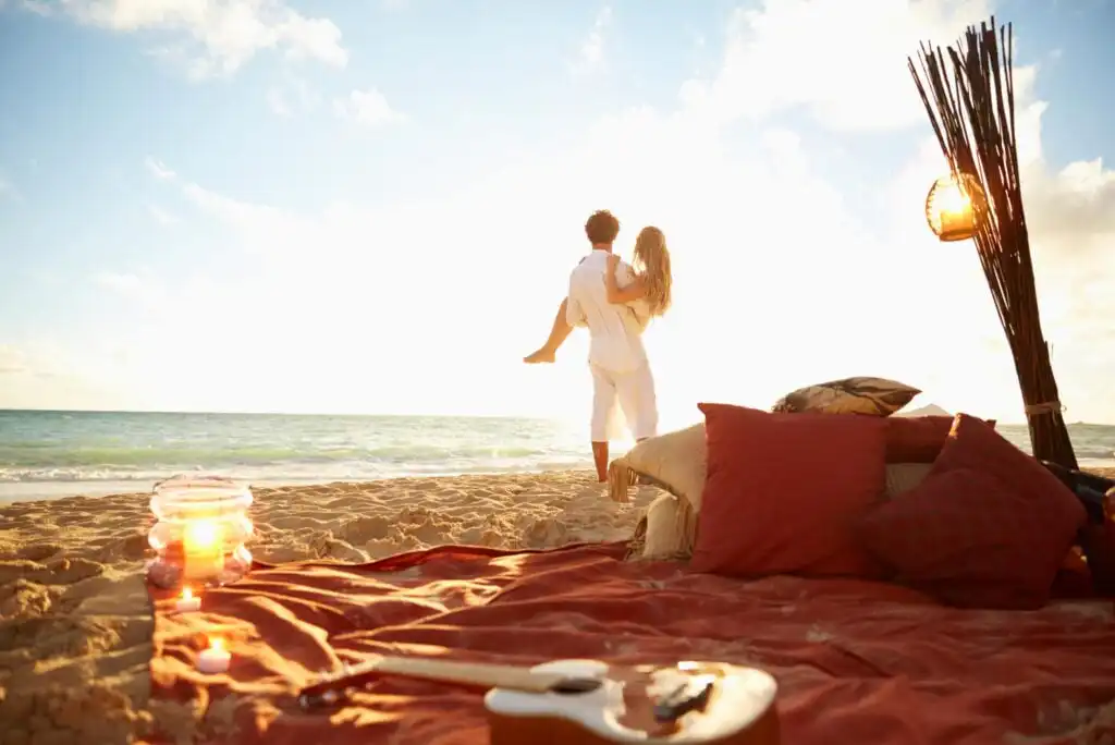 A man carries a woman in his arms on a sandy beach at sunset. In the foreground, there are red pillows, a blanket, a candle, and a guitar, creating a cozy, romantic atmosphere. The ocean and sky are in the background.