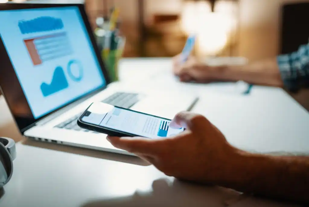 A person sits at a desk using a smartphone while taking notes with a pen, with a laptop displaying charts and graphs in the background. The workspace is well lit and organized.