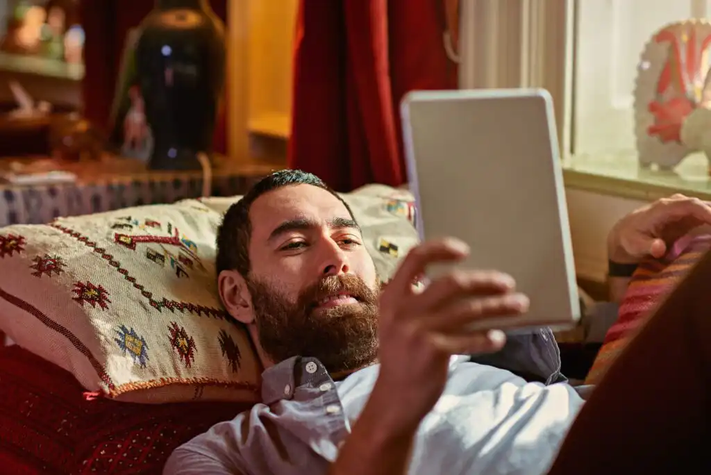 A man with a beard reclines on a couch, smiling as he holds a tablet. The room has warm lighting, patterned pillows, and decorative items in the background.