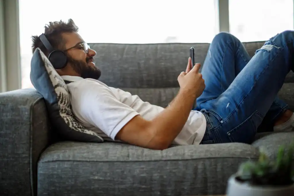 A man wearing headphones and glasses is lying on a gray sofa, smiling while looking at his smartphone. He is dressed casually in a white shirt and blue jeans.