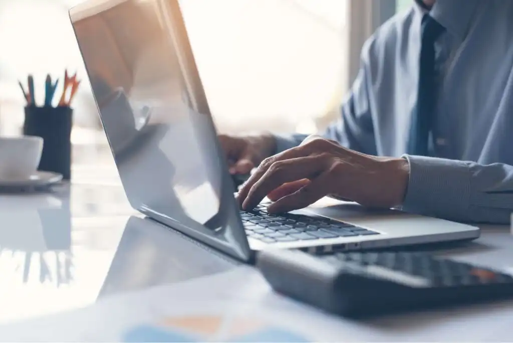 A person in a dress shirt types on a laptop at a desk, with a calculator and documents nearby. Sunlight streams in from a window, creating a bright, professional workspace environment.
