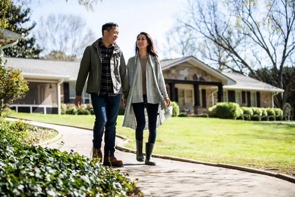 A couple walks hand in hand on a sidewalk in a residential neighborhood, smiling and talking. They wear casual clothes and boots, with houses and green lawns in the background under a clear sky.