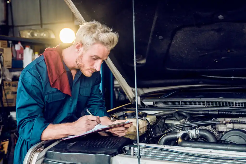 A mechanic in a blue jumpsuit writes on a clipboard while inspecting a car engine with the hood open inside a garage.