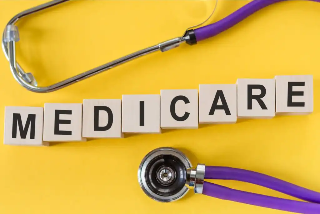 Wooden blocks spelling MEDICARE are arranged on a yellow surface, with a purple stethoscope positioned above and below the blocks.