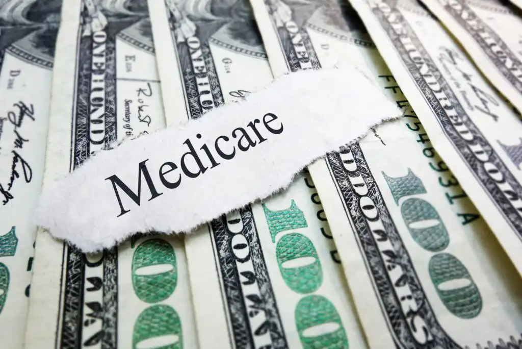 A torn piece of paper with the word Medicare sits on top of several overlapping one hundred dollar bills.