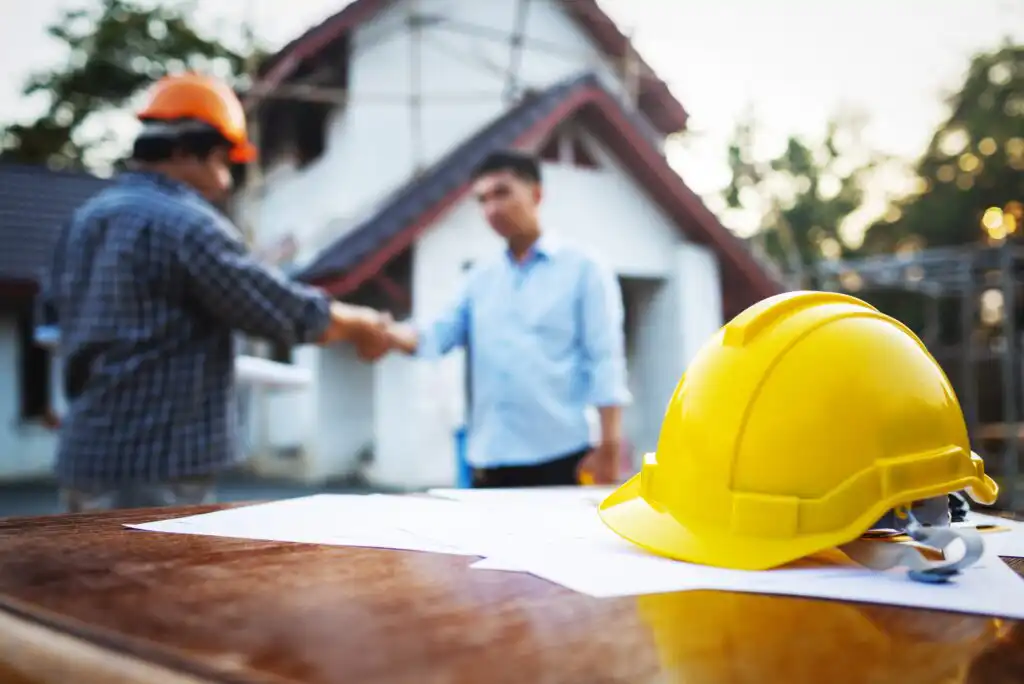 A yellow construction helmet and blueprints rest on a table, while two people in safety gear shake hands in front of a house under construction in the background.
