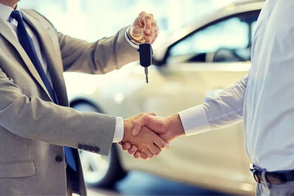 A person in a suit hands over car keys to another person while shaking hands, with a blurred car in the background, suggesting a successful car sale or purchase.