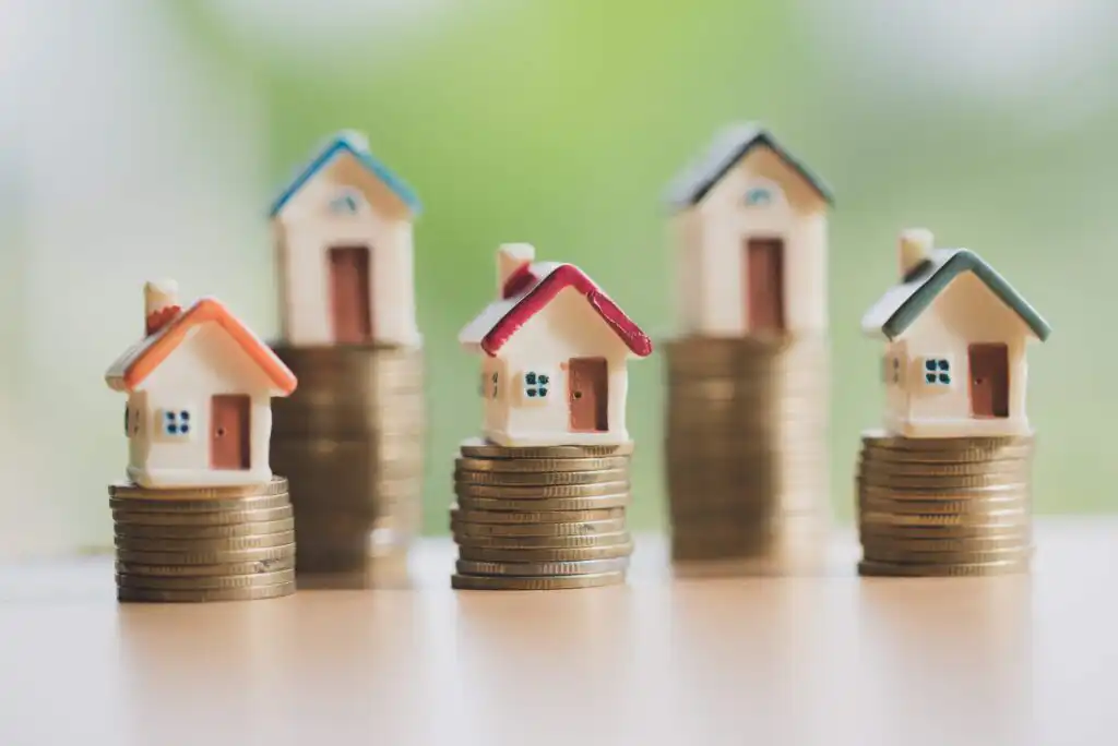 Four small model houses sit atop stacks of coins, symbolizing real estate investment, property value, or home financing, with a soft green and beige blurred background.
