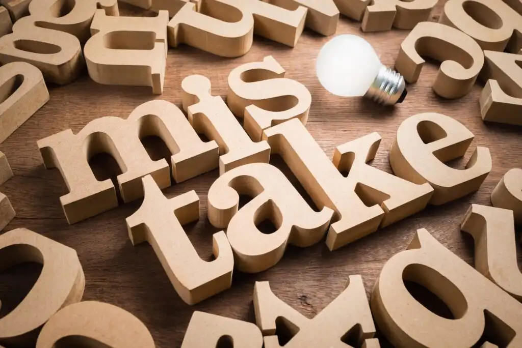 Wooden block letters spell out the word “mistake” on a wooden surface, surrounded by other scattered letters. A light bulb sits near the top right corner of the image, symbolizing an idea or realization.