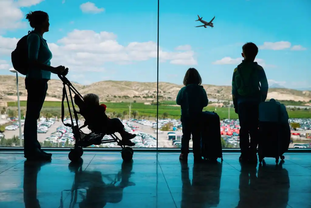 Silhouette of a woman with a child in a stroller and two children with suitcases looking out an airport window at a plane flying in the blue sky above a parking lot and hills.