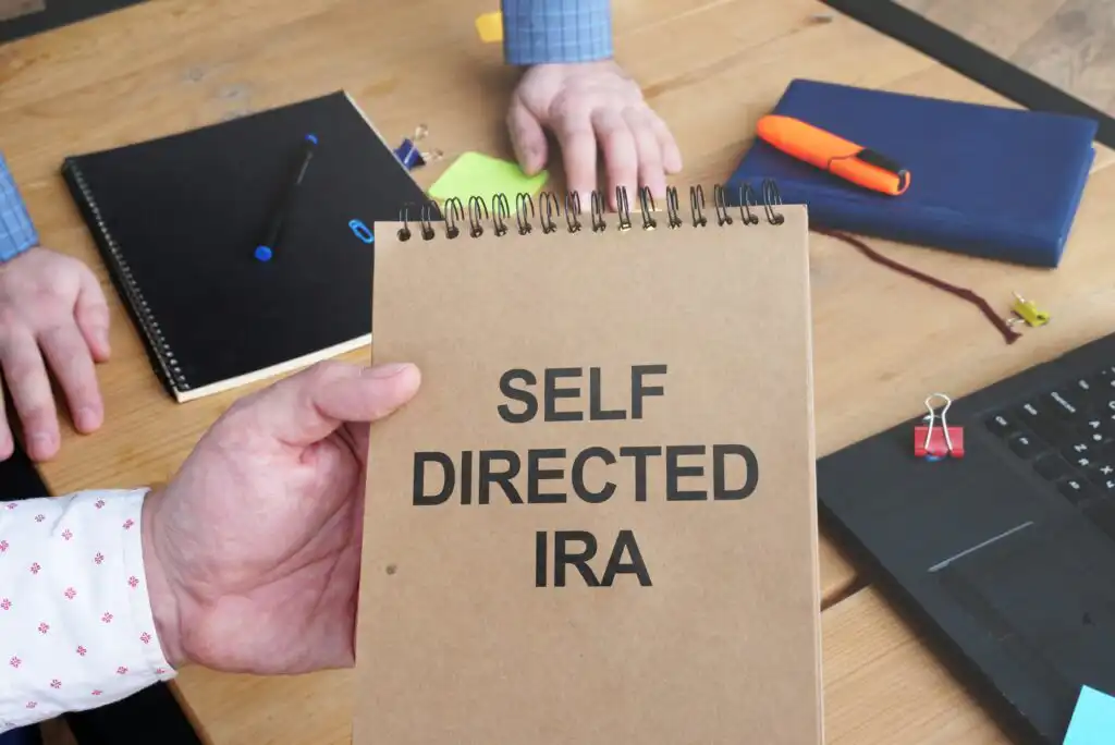 A person holds a brown spiral notebook labeled SELF DIRECTED IRA at a desk with notebooks, pens, a laptop, and office supplies in the background.
