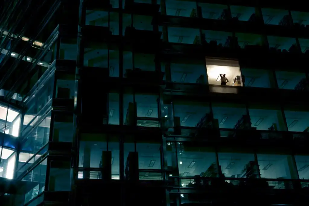 A dark office building at night with one brightly lit window, showing the silhouette of a person standing and working at a desk, surrounded by shelves and office furniture.