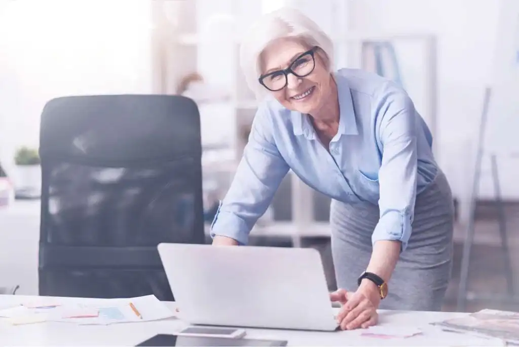 An older woman with short gray hair and glasses smiles while leaning over a desk with a laptop in an office setting, wearing a light blue blouse and gray skirt.