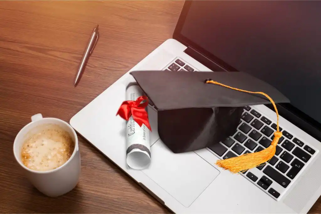 A graduation cap, diploma with a red ribbon, and cup of coffee are placed on a laptop keyboard on a wooden desk, next to a pen.