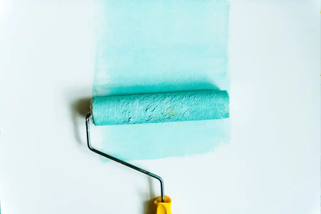 A paint roller with a yellow handle is applying light blue paint onto a white surface, leaving a fresh, smooth layer of color.