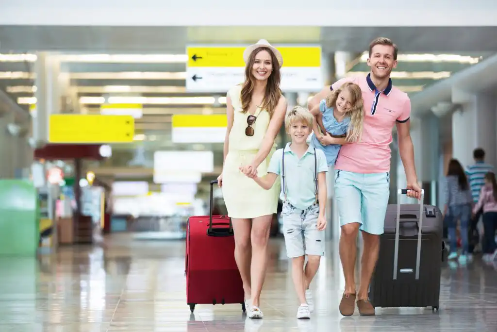 A smiling family of four with suitcases walks through an airport. The parents hold hands, the mother has a red suitcase, the father carries a gray one, and the children walk beside them, with the little girl being carried piggyback.