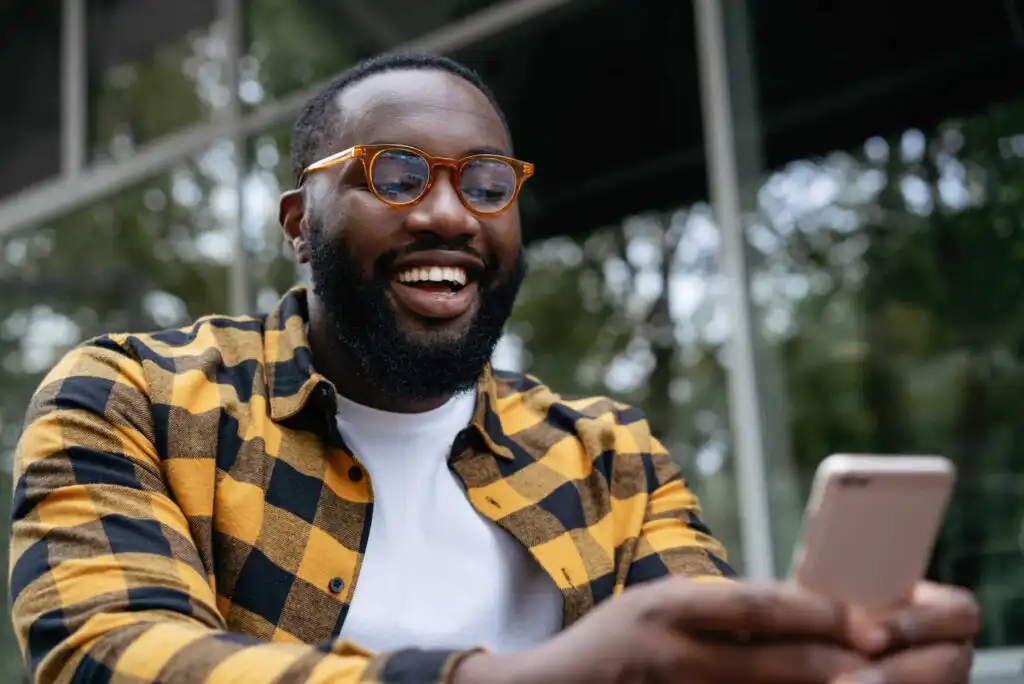 A smiling man with a beard and glasses, wearing a yellow and black plaid shirt, is sitting outdoors and holding a smartphone, looking at the screen.