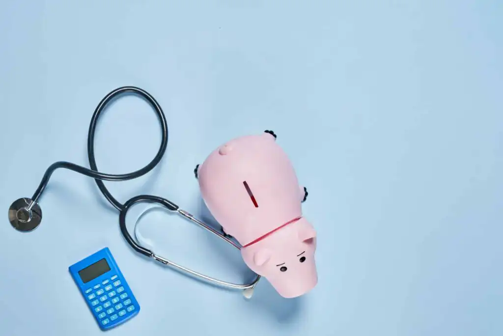 A pink piggy bank sits on a light blue surface with a stethoscope wrapped around it and a blue calculator nearby, symbolizing financial health or healthcare costs.